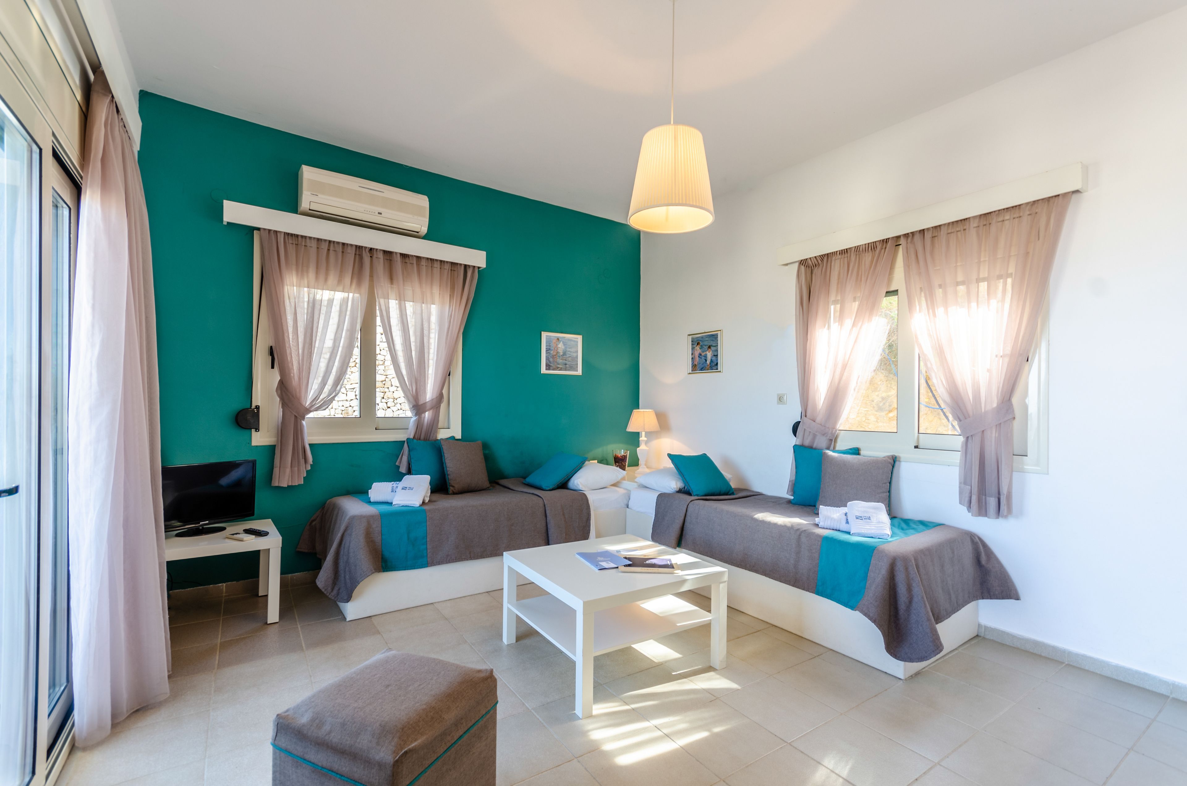 A modern room with teal walls, two beds, a white coffee table, a TV, and an air conditioning unit.