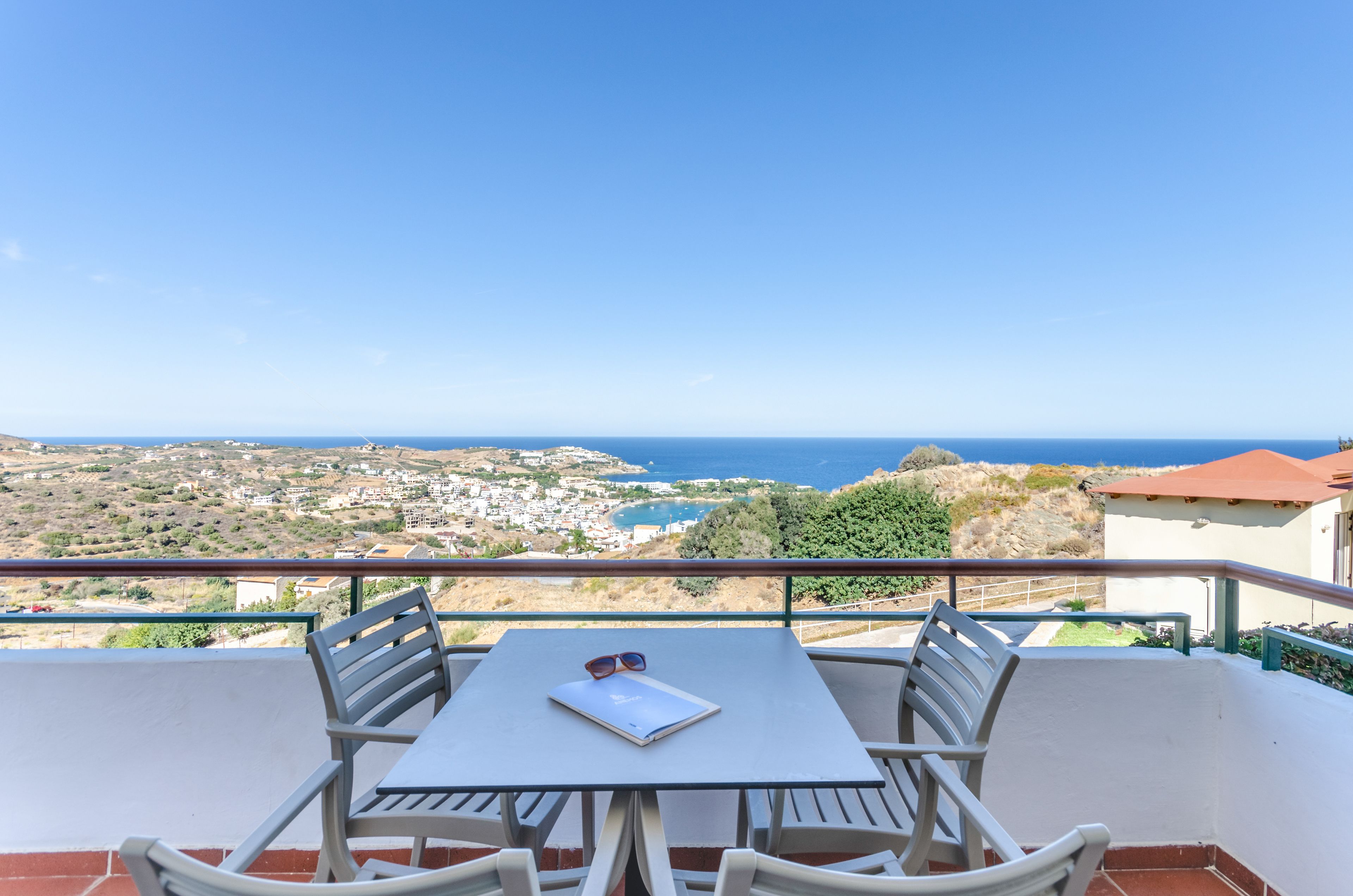 A balcony with a table and chairs, overlooking a coastal town and the sea.