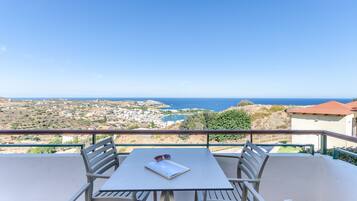 A balcony with a table and chairs, overlooking a coastal town and the sea.