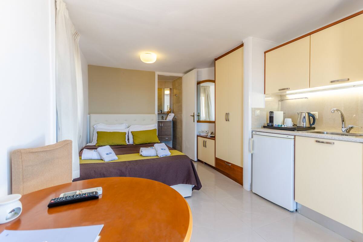 Une chambre d’hôtel comprenant un lit, une petite table avec une télécommande, un coin cuisine avec un évier et une machine à café, et un miroir au-dessus de l’évier.
