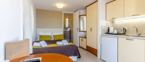 A hotel room with a bed, a small table with a remote control, a kitchen area with a sink and a coffee maker, and a mirror above the sink.