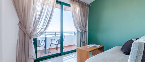 A hotel room with a bed, a small wooden table, a chair, and a balcony with a view.