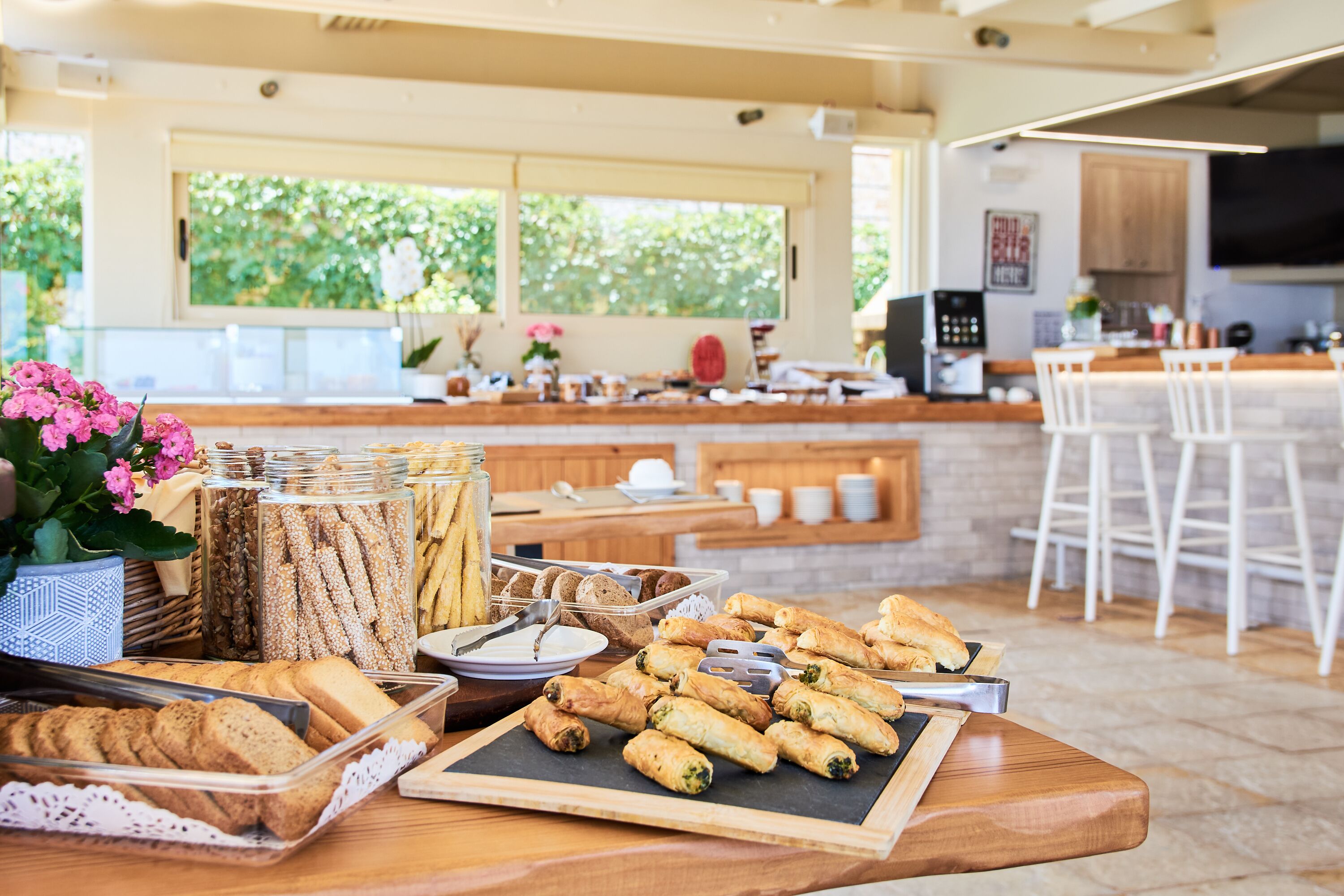 Een ontbijtbuffet met diverse gebakjes en brood, een koffiehoek, en uitzicht op het omliggende landschap door grote ramen.