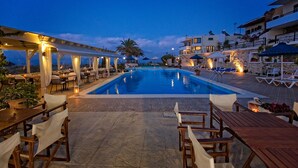 A poolside area with a pool, lounge chairs, and tables, surrounded by buildings and a palm tree. - Pela Mare Hotel (Malevizi)
