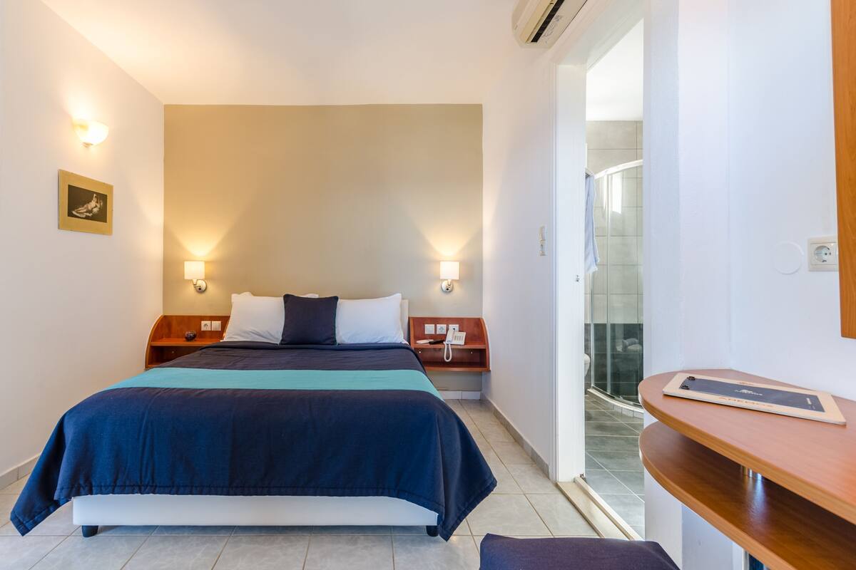 Une chambre d’hôtel avec un lit, des lampes de chevet, un téléphone sur la table de chevet, une tablette posée sur une petite table en bois, et un espace douche visible à travers une porte en verre.