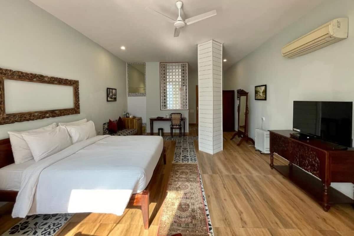 A bedroom with a large bed, a ceiling fan, an air conditioning unit, a television, and a desk with a chair.