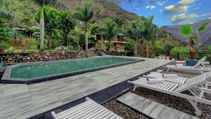 A pool area with lounge chairs, a stone wall, and a view of a mountainous landscape. - Raga on the Ganges (Narendranagar)