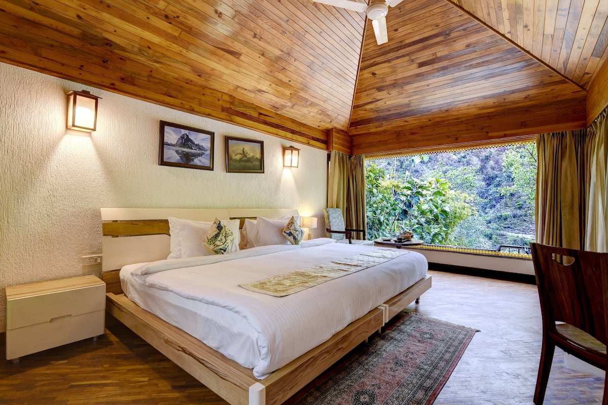 A bedroom with a wooden ceiling, a large bed with white bedding, a nightstand, a lamp, and a view of greenery through a window.