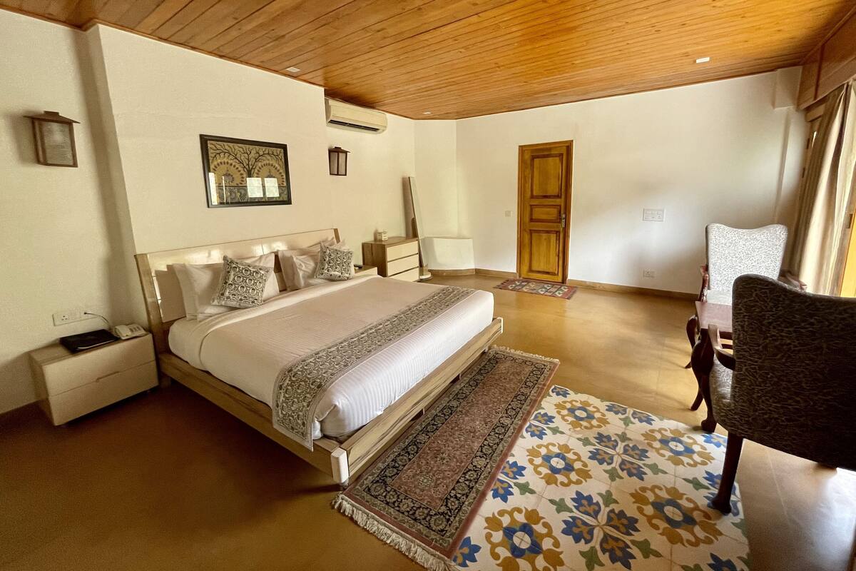 A bedroom with a large bed, bedside tables, a nightstand, a wooden ceiling, a door, a chair, and a patterned rug.