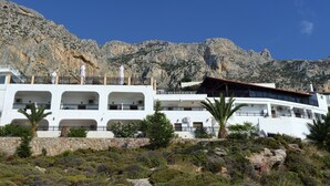 Un edificio blanco con varios balcones, rodeado de terreno rocoso y vegetación escasa.