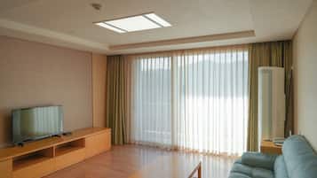 A modern living room with a flat-screen TV, wooden furniture, a sofa, and large windows with sheer curtains.