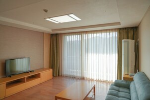 Una sala de estar moderna con un televisor de pantalla plana, muebles de madera, un sofá y ventanas grandes con cortinas translúcidas.