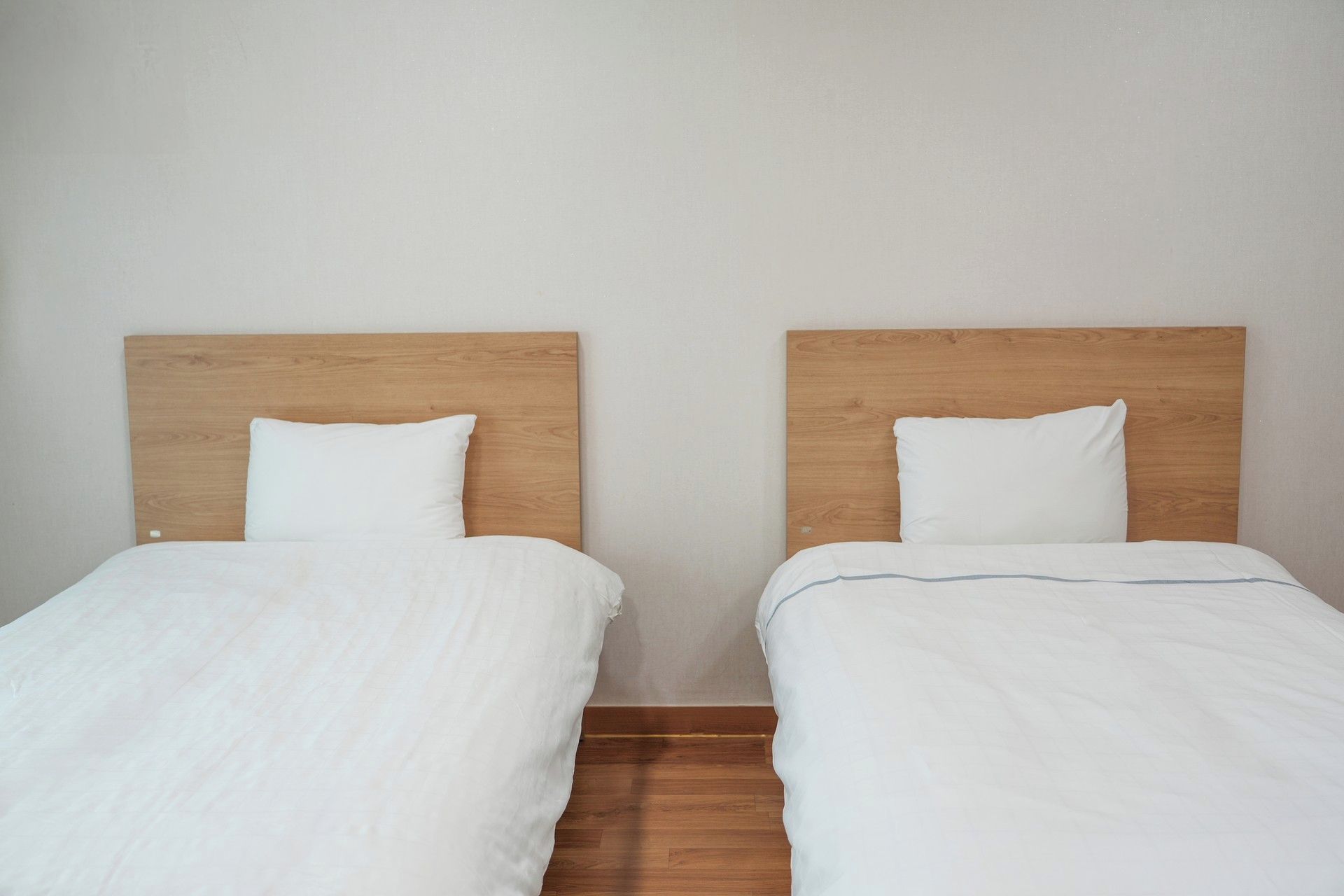 Dos camas individuales con cabeceros de madera y ropa de cama blanca en una habitación con pared lisa y suelo de madera.