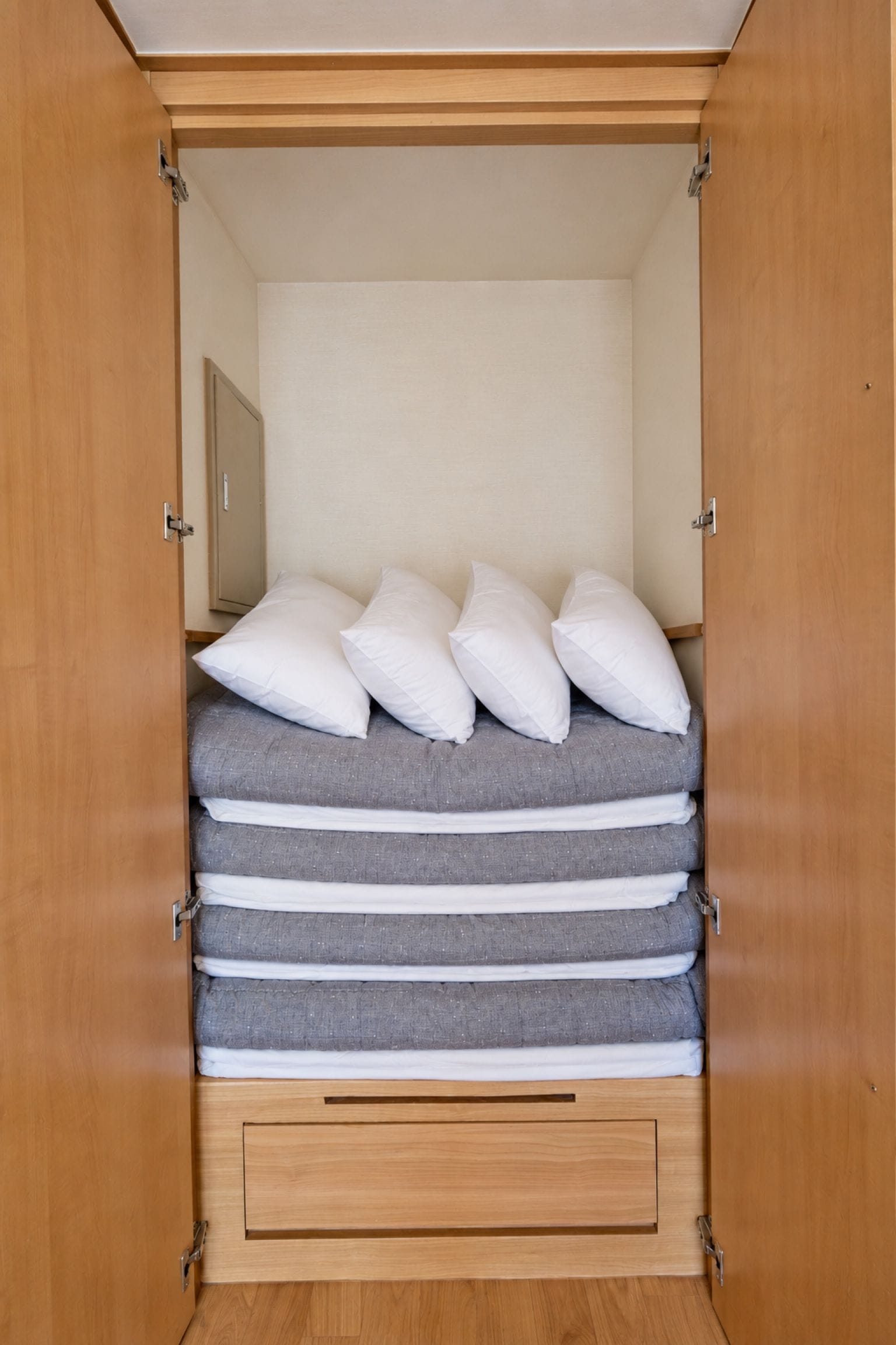 A wooden wardrobe with a bed inside, featuring multiple pillows and a grey mattress.