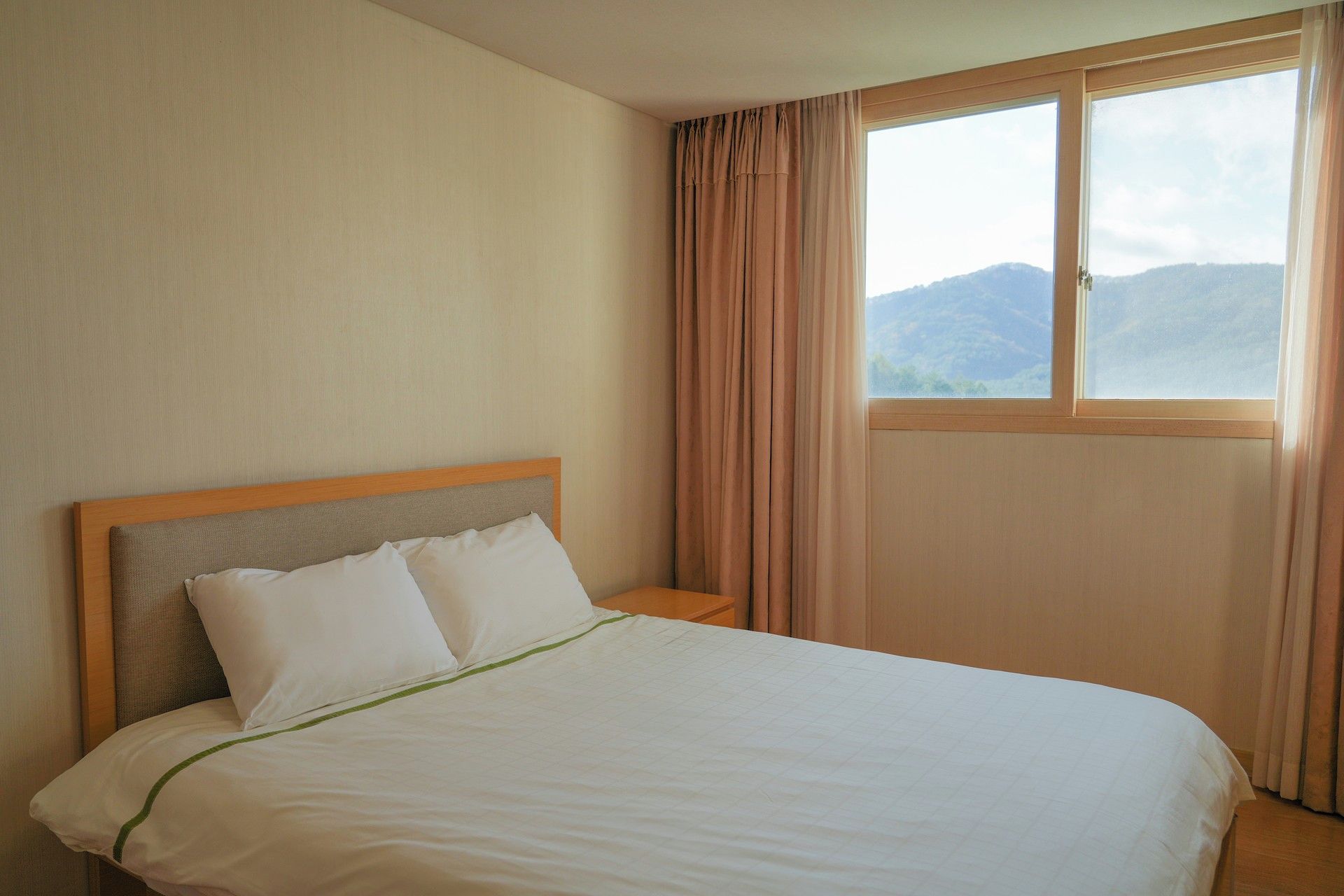 A hotel room with a bed, a nightstand, a window with curtains, and a view of mountains.