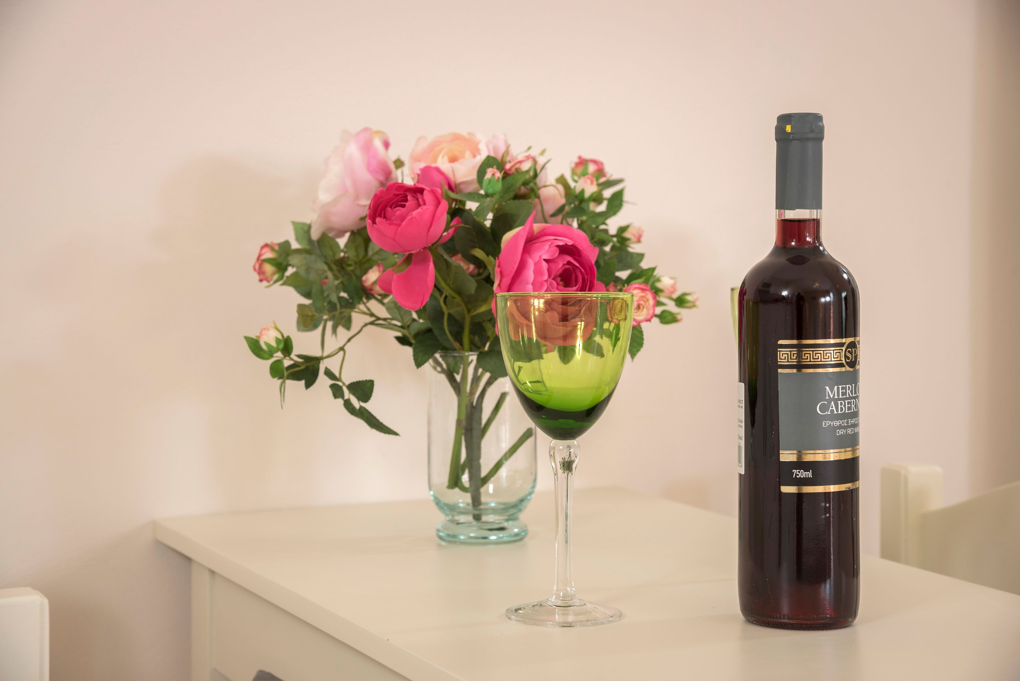Una botella de Merlot Cabernet y una copa de vino sobre una mesa con un jarrón de rosas rosadas.