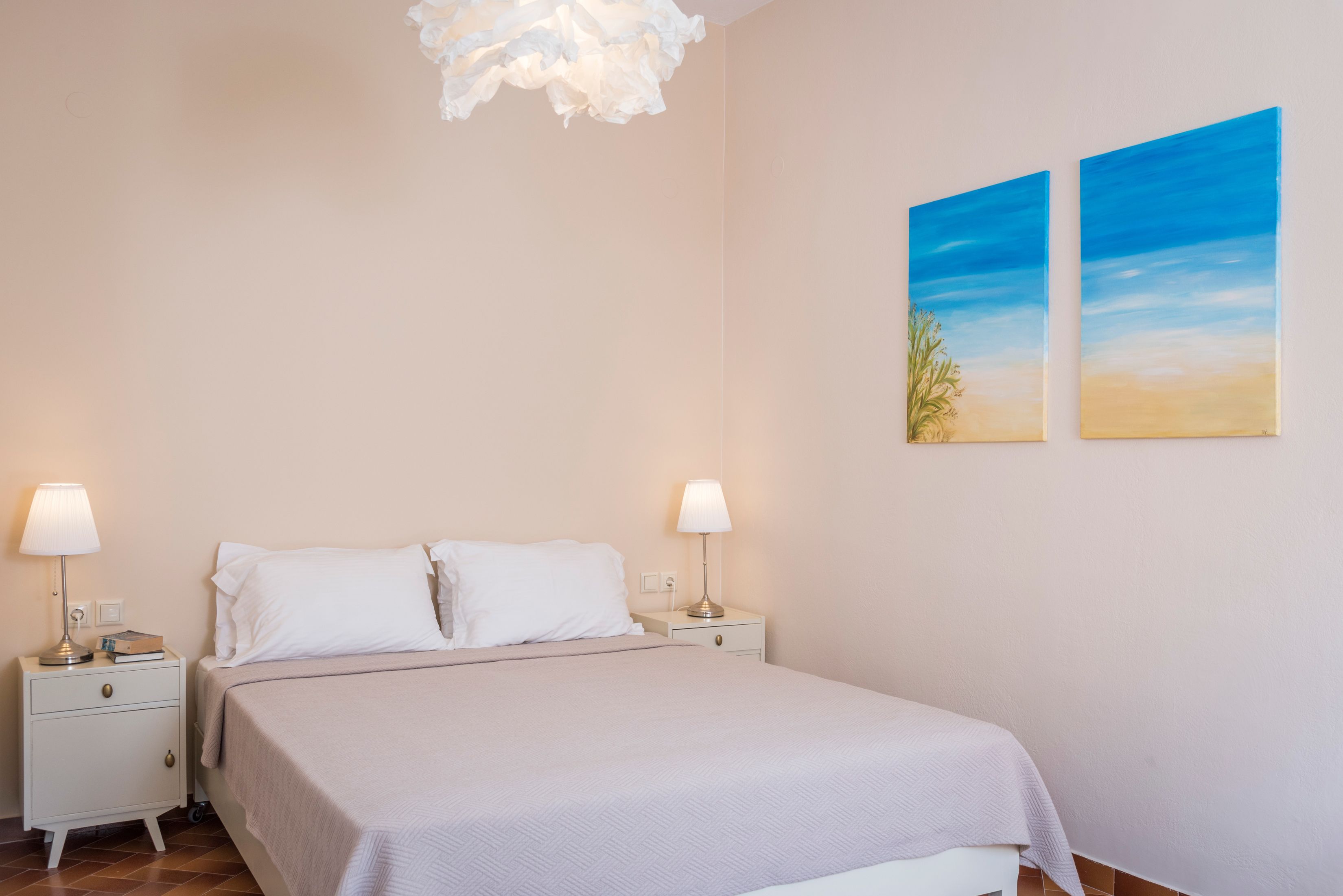 A bedroom with a bed, two bedside tables, two wall-mounted lamps, and two paintings.