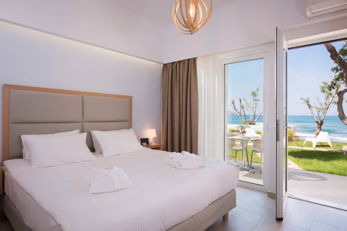 A hotel room with a large bed, a nightstand, a lamp, and a view of the ocean through a sliding glass door.