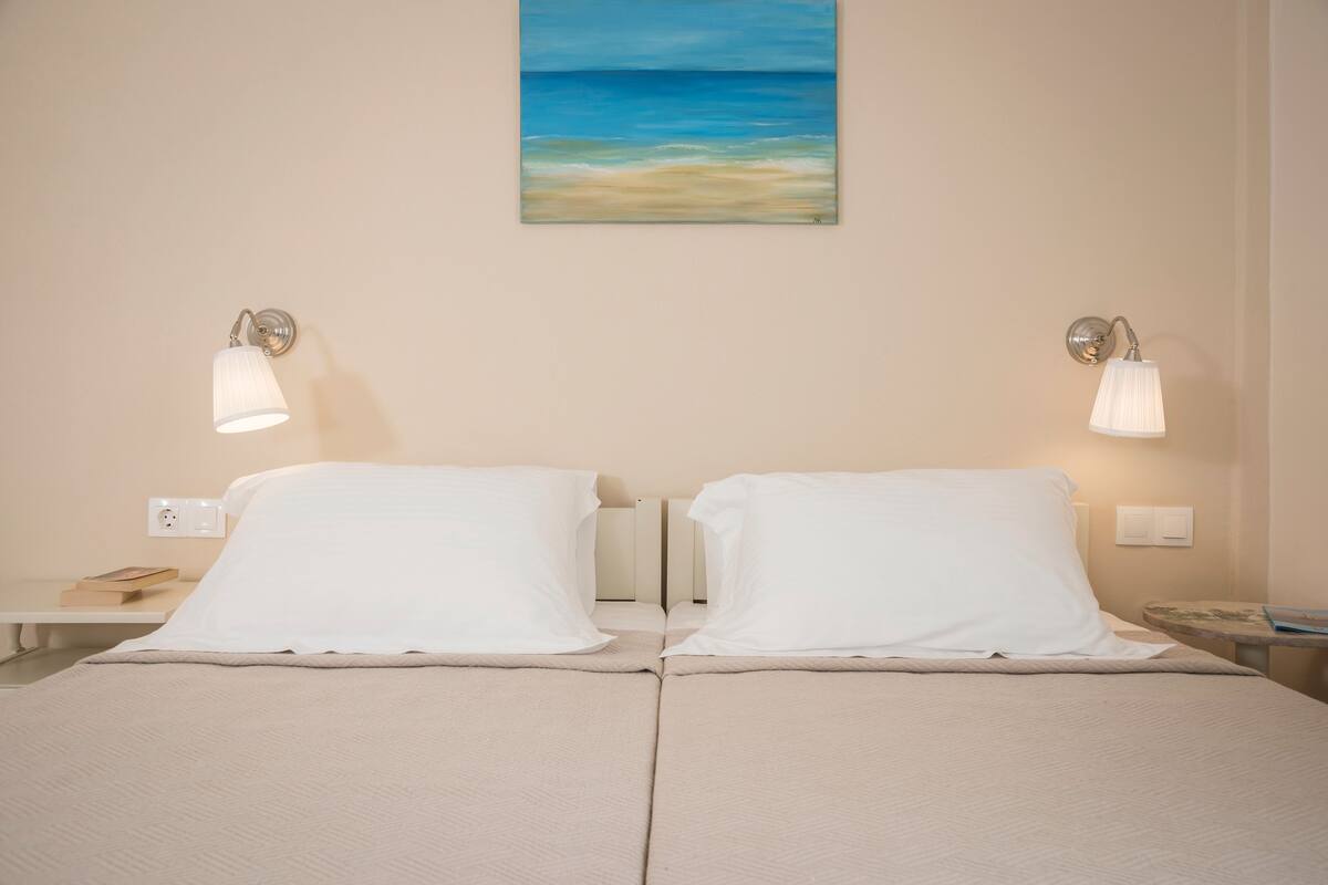A double bed with white linens, two bedside tables, a wall-mounted painting of a beach scene, and two wall-mounted lamps.