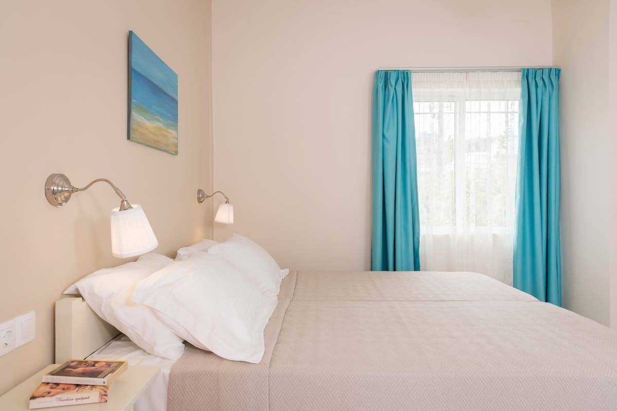 A neatly made bed with white linens, a bedside table with a lamp, and a window with teal curtains.