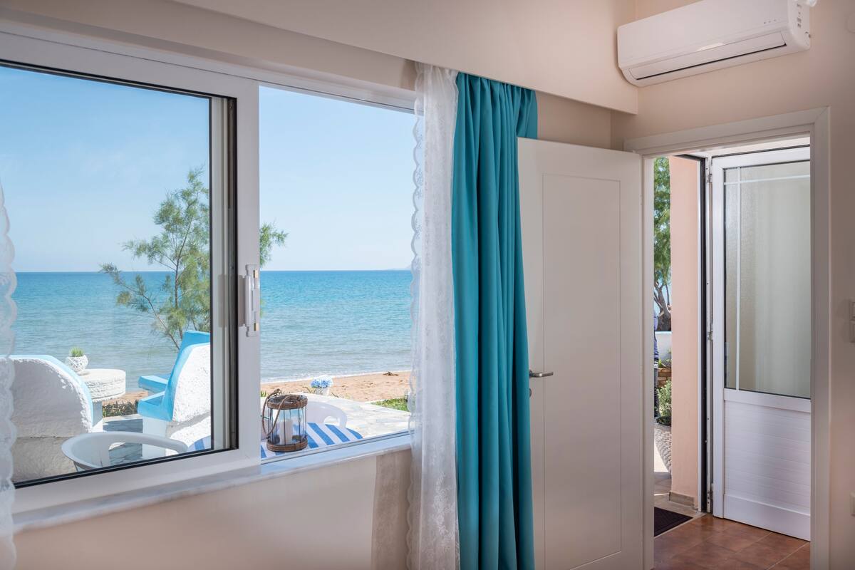 A room with a large window overlooking a beach, a white door, and a view of the sea.
