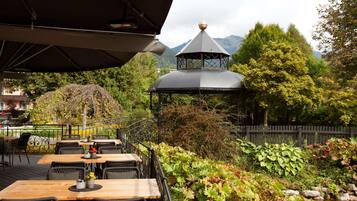 An outdoor dining area with tables and chairs, surrounded by lush greenery and a mountain backdrop.