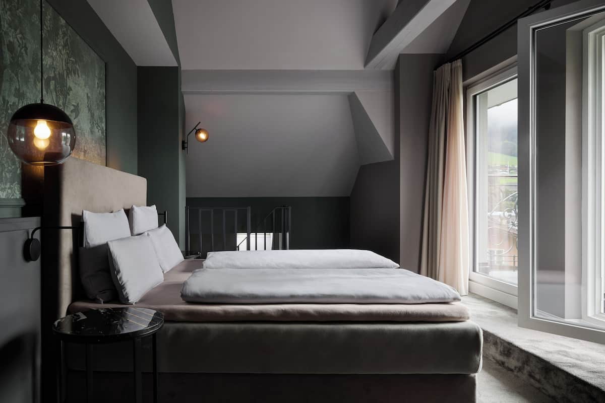 Un dormitorio moderno con una cama grande, una mesita, una lámpara de pared y una ventana con cortinas.