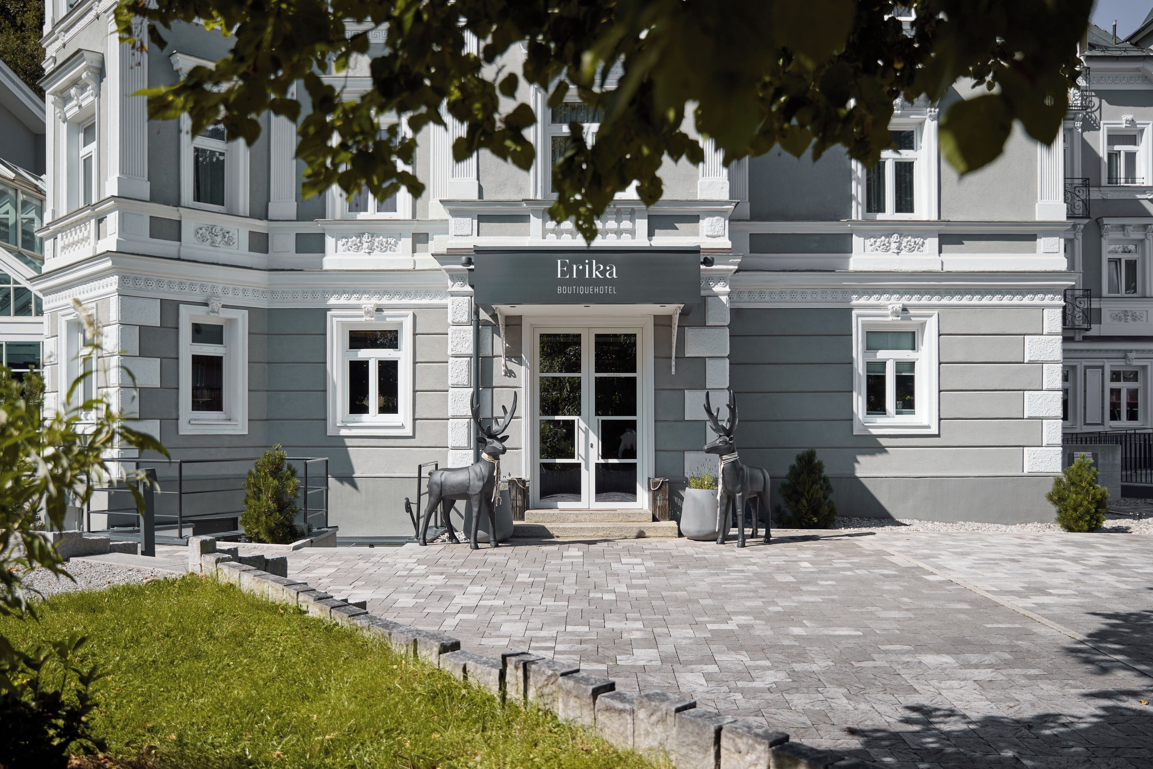En elegant hotellentrén med en skylt som visar texten ”Erika Boutique Hotel”, en asfalterad uppfart och två statyer föreställande rådjur.