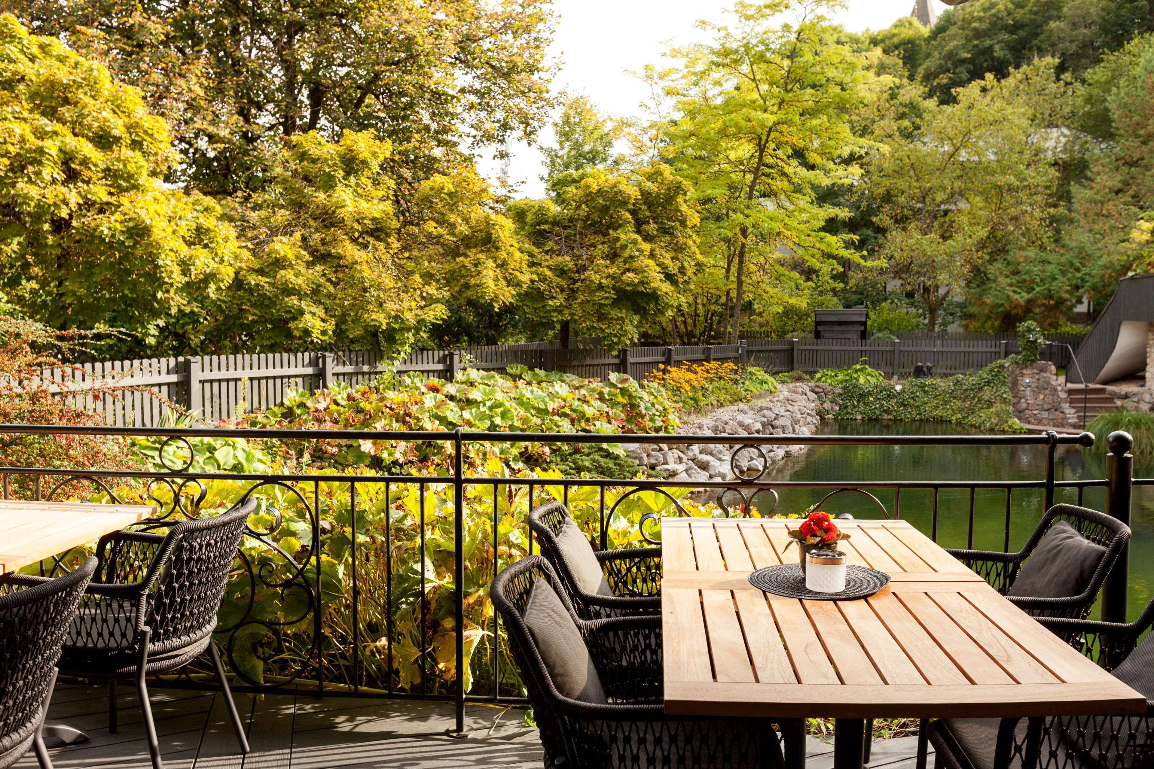 An outdoor patio with a wooden table and wicker chairs, surrounded by lush greenery and a stone wall.