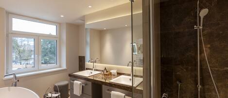 Un baño moderno con una bañera grande, dos lavamanos, una cabina de ducha de cristal y una ventana que proporciona luz natural.