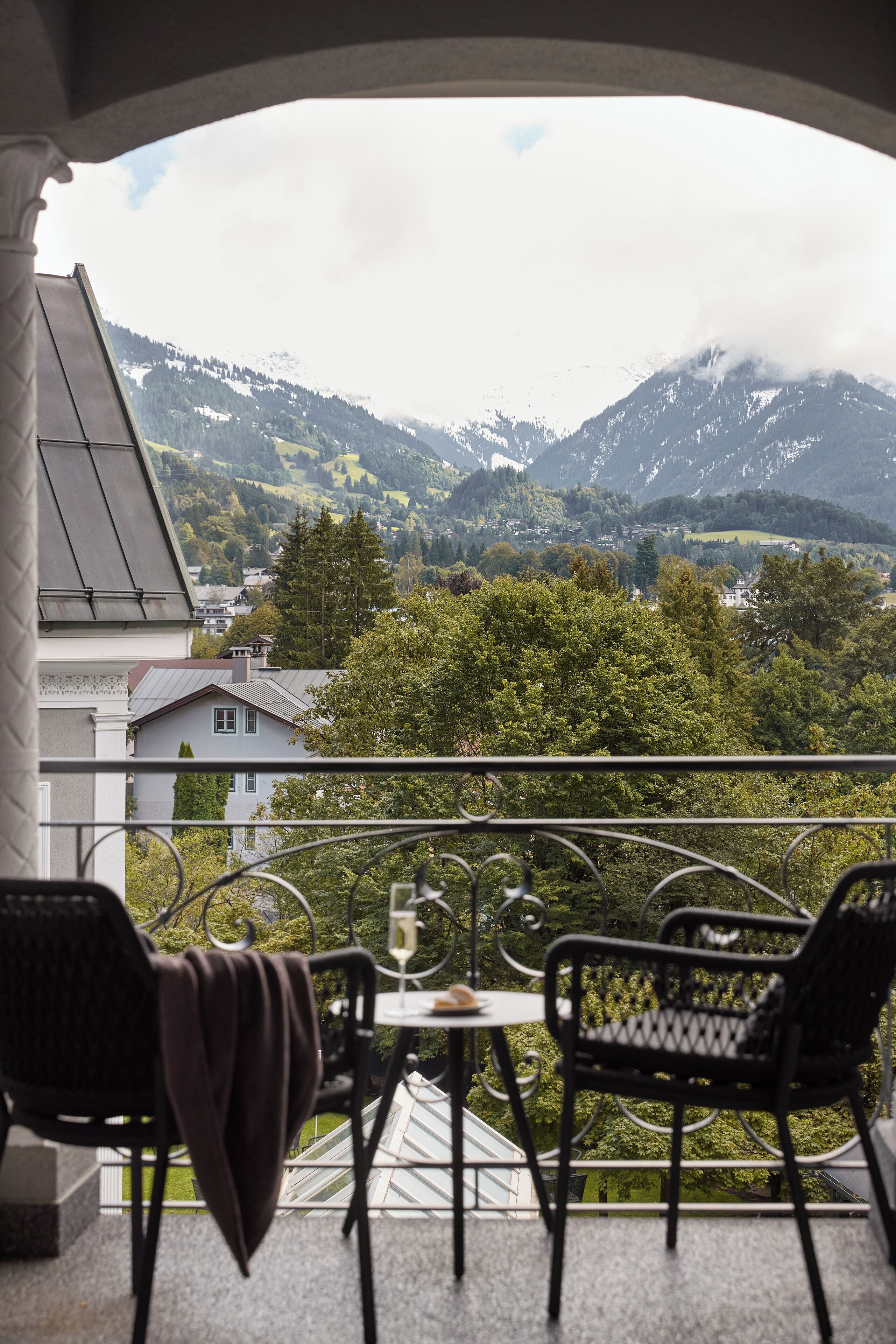Ein Balkon mit zwei Stühlen und einem Tisch, der auf eine gebirgige Landschaft mit Häusern und viel Grün blickt.