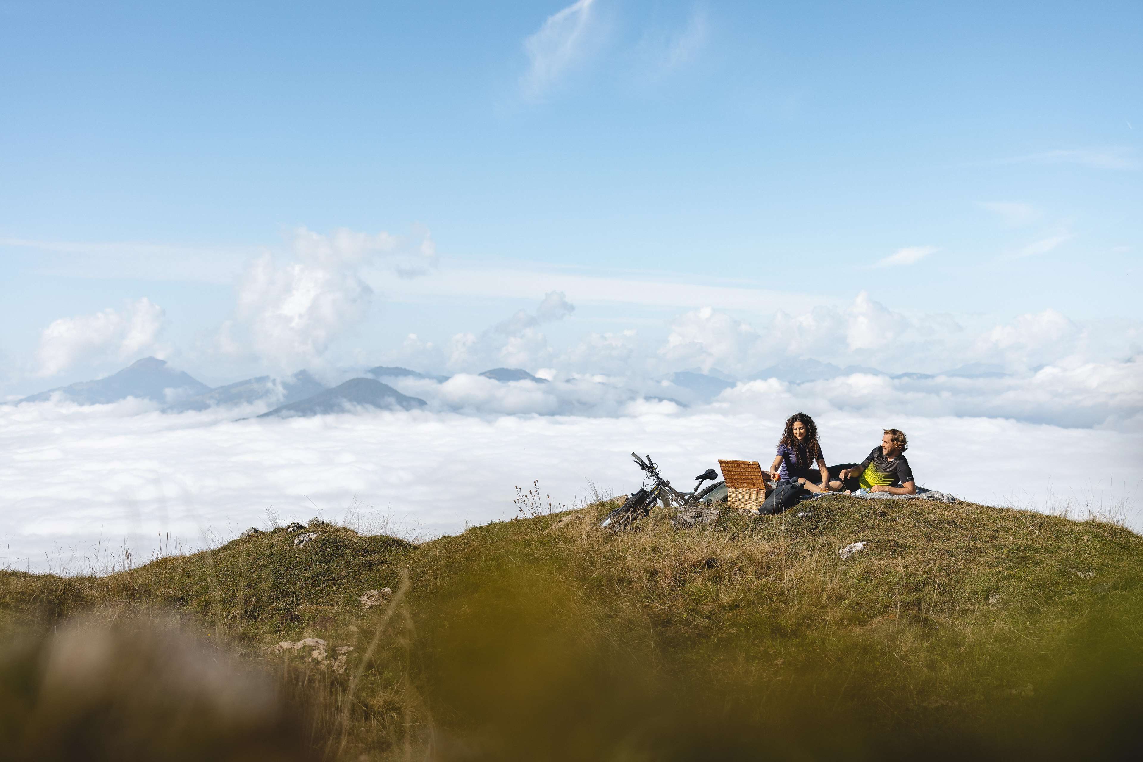 Dos personas sentadas en una colina cubierta de hierba, con una montaña y nubes al fondo.