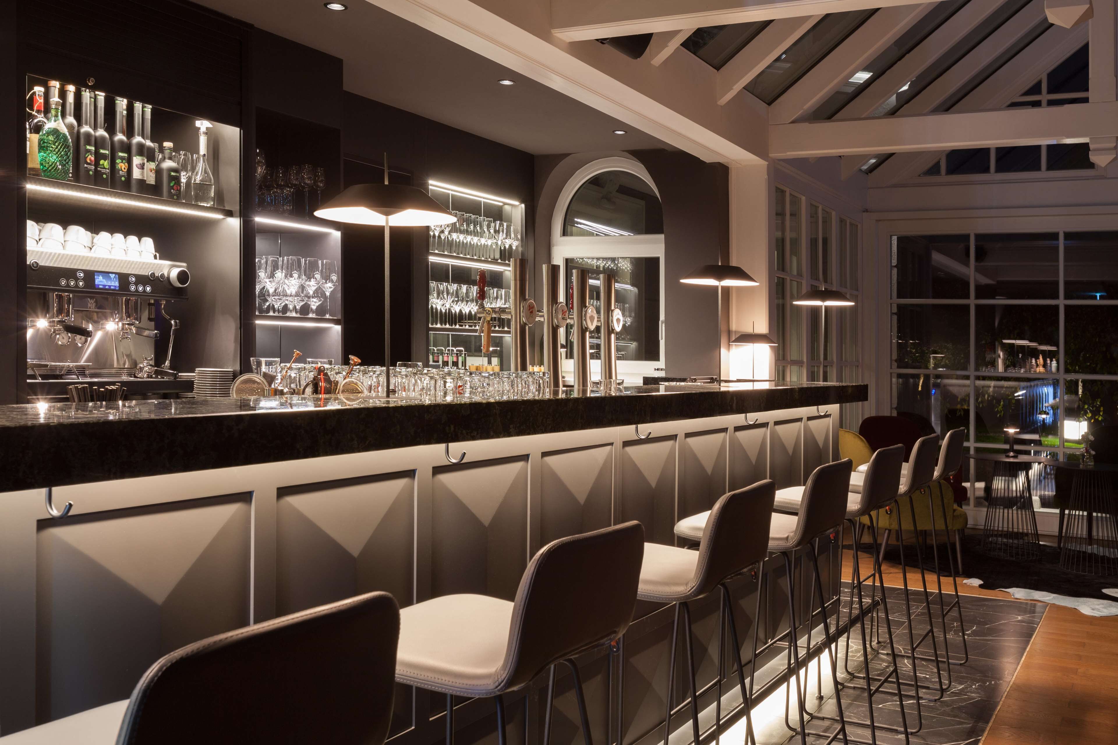 A bar area with a long counter, multiple stools, and shelves stocked with bottles and glasses.