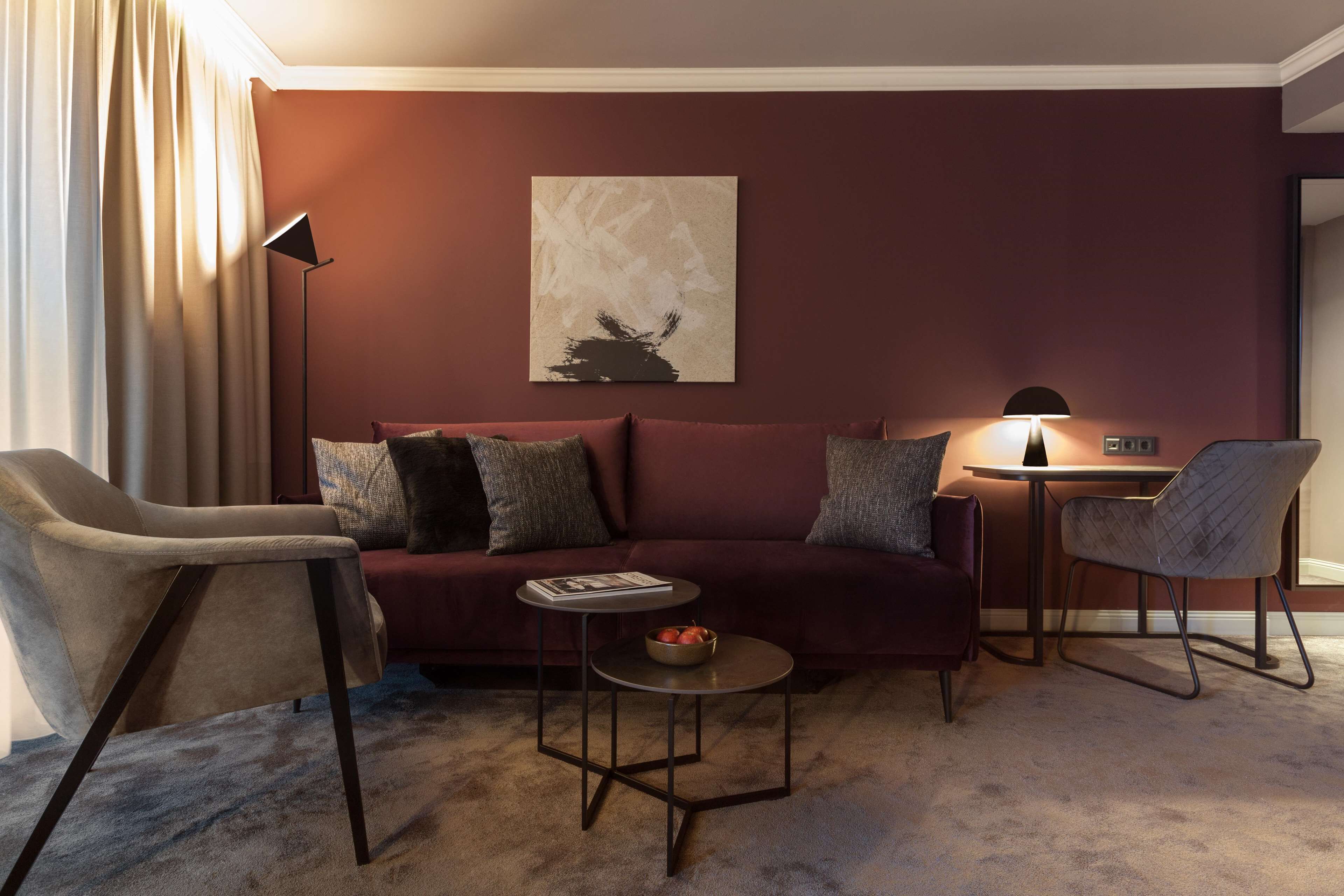 A modern living room with a dark red wall, a beige sofa with multiple cushions, a round coffee table with a bowl of fruit, a floor lamp, and a side table with a lamp.