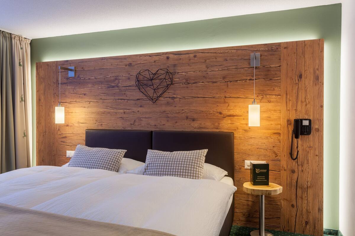 Una habitación de hotel con una cama grande, un cabecero de madera, una decoración en forma de corazón en la pared, lámparas de noche y un teléfono sobre una mesita.