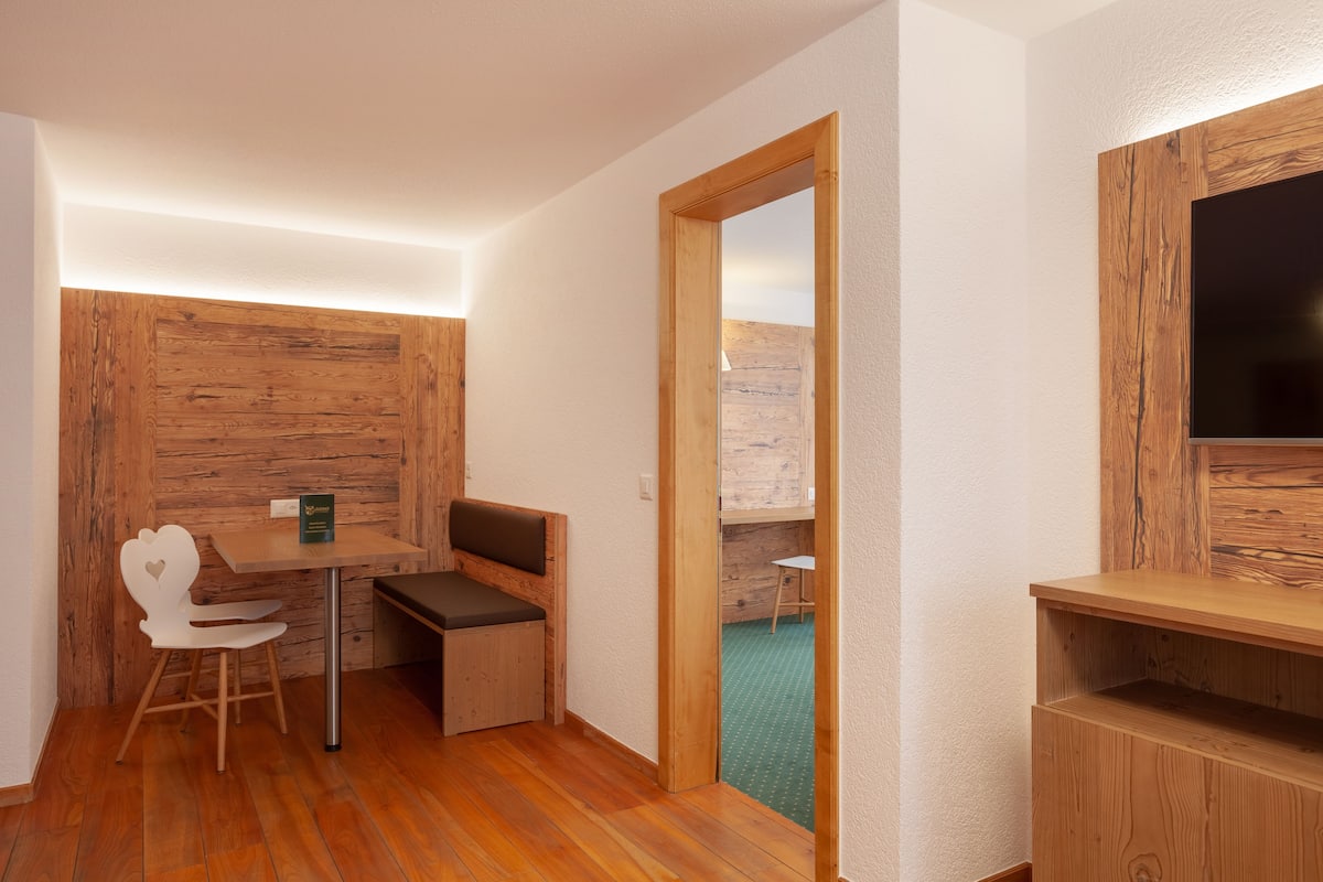 Una habitación compacta con una pared de madera, una pequeña mesa con un banco, una silla blanca y un televisor fijado a la pared.