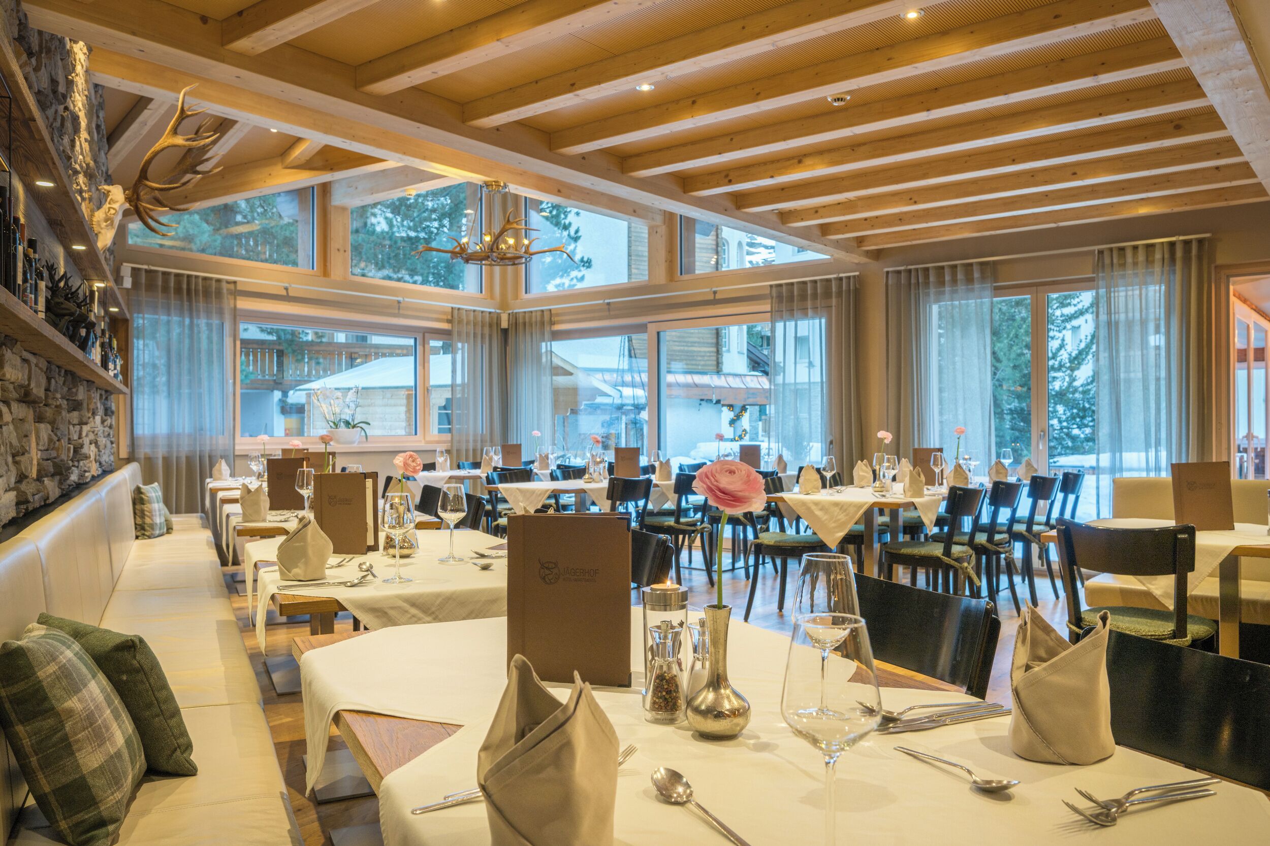 A restaurant with wooden beams, large windows, and a stone wall with antlers. Tables are set with white tablecloths, silverware, and a vase with a flower.