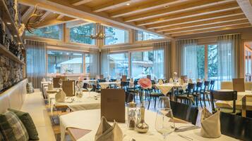 A restaurant with wooden beams, large windows, and a stone wall with antlers. Tables are set with white tablecloths, silverware, and a vase with a flower.