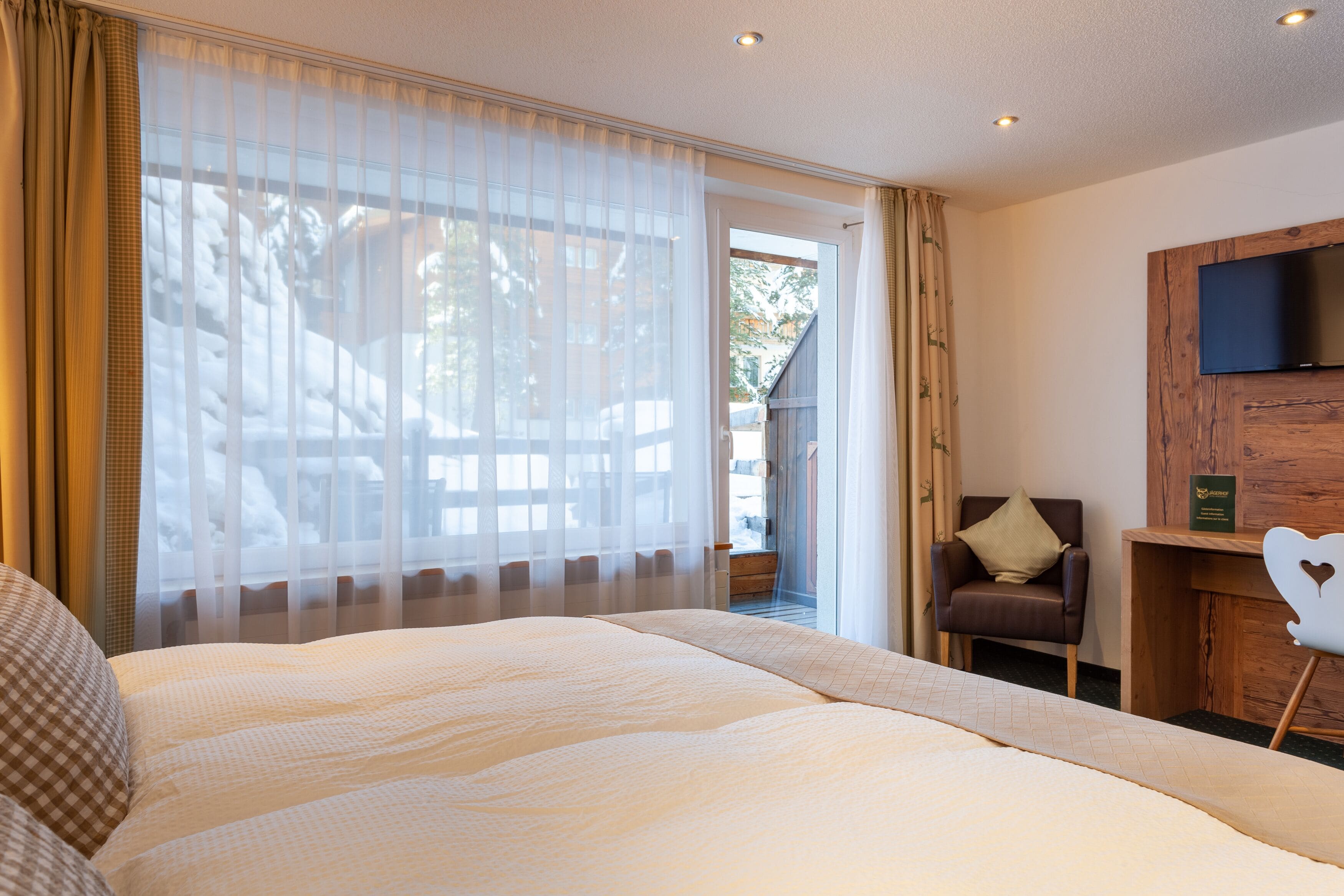 Habitación de hotel con una cama grande, una silla, un escritorio con televisor de pantalla plana y vistas a través de una ventana con cortinas translúcidas.