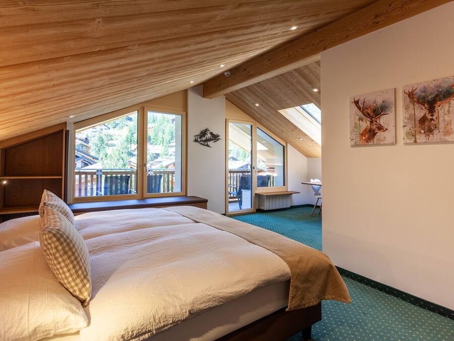 A bedroom with a large bed, wooden ceiling beams, a view of the outdoors, and two deer paintings on the wall.