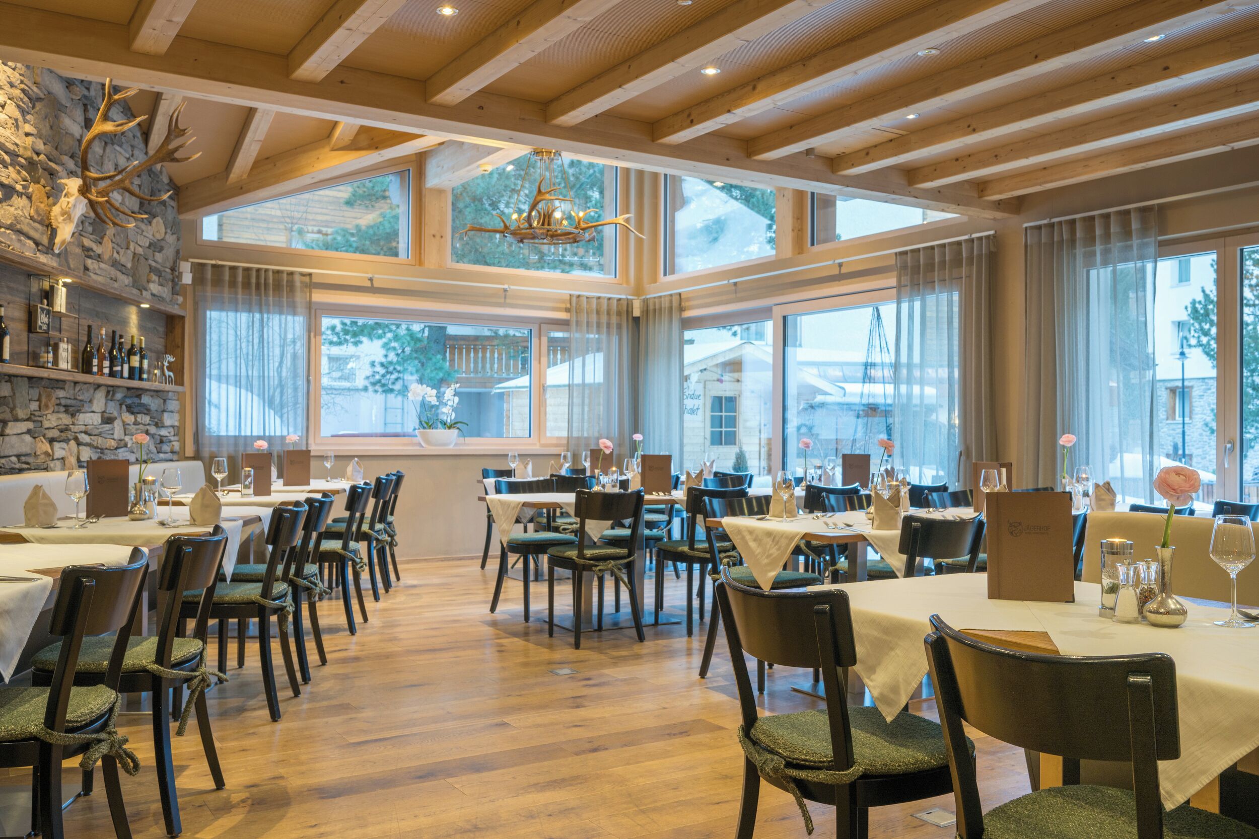 A restaurant with wooden beams, stone walls, and large windows. Tables are set with chairs and wine glasses.