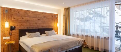 A hotel room with a large bed, wooden headboard, a small side table, and a window with sheer curtains.