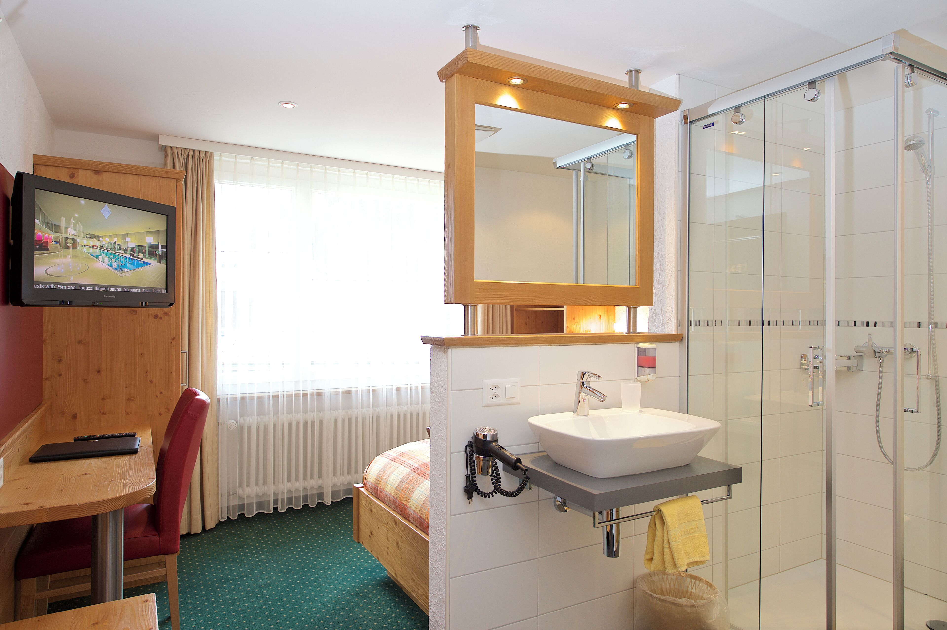 A hotel room with a wooden desk, a bed with a plaid blanket, a bathroom with a glass shower, and a TV mounted on the wall.
