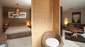 A modern bedroom with a bed, bedside lamps, a wicker chair with a cushion, a wooden partition, and a sofa in a living area. - Les Terrasses de Castelnau (Castelnau-le-Lez)