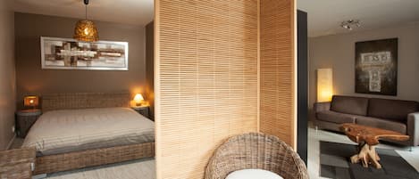 A modern bedroom with a bed, bedside lamps, a wicker chair with a cushion, a wooden partition, and a sofa in a living area.