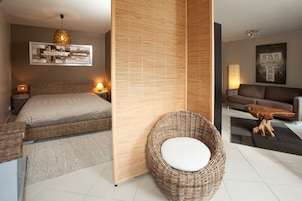 Un dormitorio moderno con una cama, lámparas de noche, una silla de mimbre con cojín, una celosía de madera y un sofá en una zona de estar.