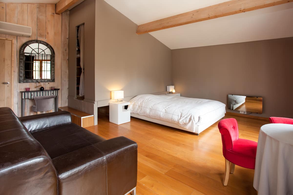 A bedroom with a bed, a red chair, a white table, a wooden floor, and a window with a mirror.