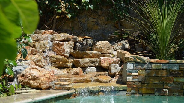 A pool with a waterfall feature, surrounded by rocks and plants. - Les Terrasses de Castelnau (Castelnau-le-Lez)
