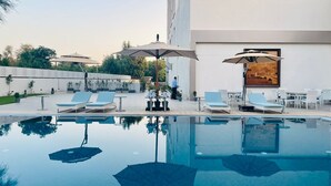 Un área de piscina moderna al aire libre con sillones de descanso y sombrillas, rodeada de un jardín ajardinado y un edificio con un mural.
