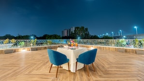 Un área de comedor al aire libre con una mesa lista para dos personas, rodeada de sillas azules, bajo un cielo nocturno con una luna visible.